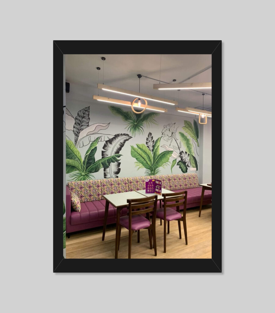 A modern café interior featuring a wall with tropical leaf murals. The seating includes a long purple cushioned bench with colorful patterned pillows and wooden chairs around light wooden tables. Pendant lights hang from the ceiling.